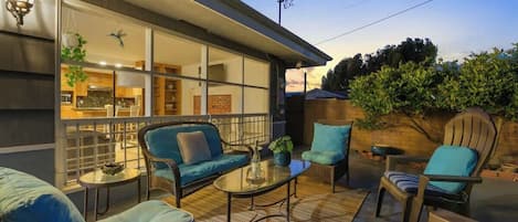 An outdoor patio area with seating, a glass door leading to a kitchen, and a view of the sky.