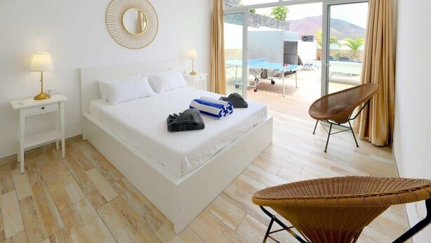 A hotel room with a large bed, a bedside table with a lamp, a wicker chair, and a view of the outdoor pool area. - Villa Brisa Summary: (Playa Blanca)