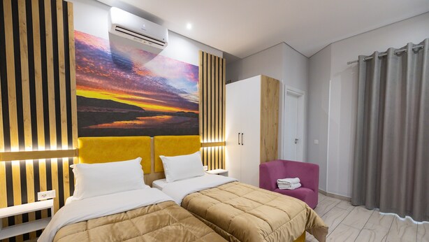 A hotel room with two beds, a large wall mural, a wooden headboard, a wardrobe, a purple armchair, and a grey curtain. - Hotel Beler (Vlorë)