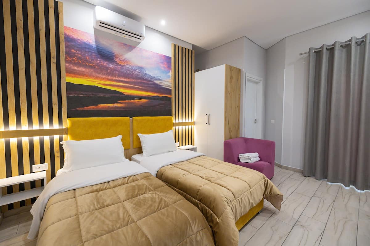 Une chambre d’hôtel avec deux lits, une grande fresque murale, une tête de lit en bois, une armoire, un fauteuil violet et un rideau gris.