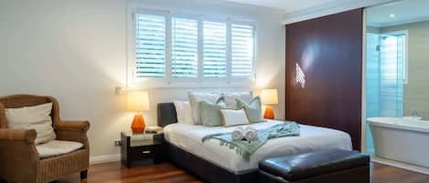 Una camera da letto con un letto, una sedia, un comodino, una lampada, una ventola a soffitto e un'ampia finestra con tende.