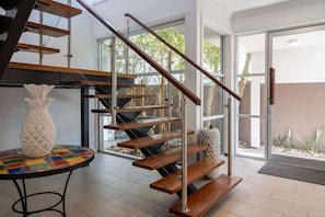 Un interior moderno con una escalera de madera, una mesa redonda con un tablero colorido y un jarrón blanco.