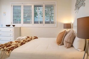 A hotel room with a bed, a bedside lamp, a window with blinds, and a dresser with a mirror.