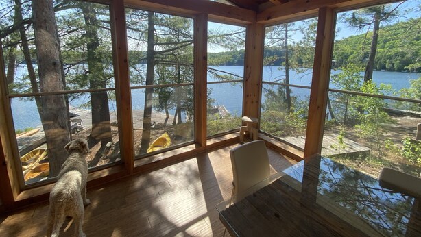 Une cabane dotée de grandes fenêtres donnant sur un lac et une forêt. À l’intérieur, on trouve une table en bois et une chaise.