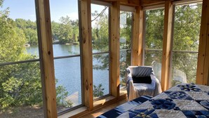 Une pièce dotée de grandes fenêtres donnant sur un lac, un lit recouvert d’une couette bleue et blanche, et une chaise placée près de la fenêtre.