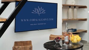 Een modern interieur met een televisie waarop een logo en website voor vakantiehuizen staan. Er is een ronde tafel met een glazen schaal vol fruit en twee wijnglazen.