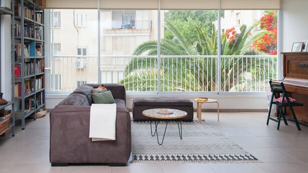 A living room with a sofa, a coffee table, a bookshelf, and a view of a balcony with plants and a building. - Spacious & Stylish 3BR Apt w Beautiful Street View (Tel Aviv)