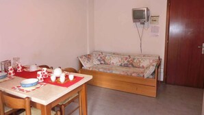 Uma área de refeições com uma mesa posta para dois, um sofá, uma televisão e uma porta.