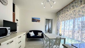 A modern kitchen with a dining area, featuring a microwave, a flat-screen TV, a dark blue sofa with two pillows, a dining table with chairs, and a large window with patterned curtains. - Holiday apartment near the beach (Bibione)
