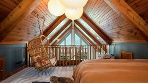 Ein Schlafzimmer mit schrägen Wänden, einer Pendelleuchte, einem Korbstuhl, einem Bett und Blick durch ein Fenster.