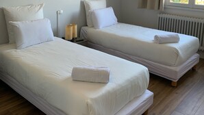 A room with two single beds, each with white bedding and pillows, a bedside table with a lamp, and a window with curtains. - Le Costellan (Dagneux)