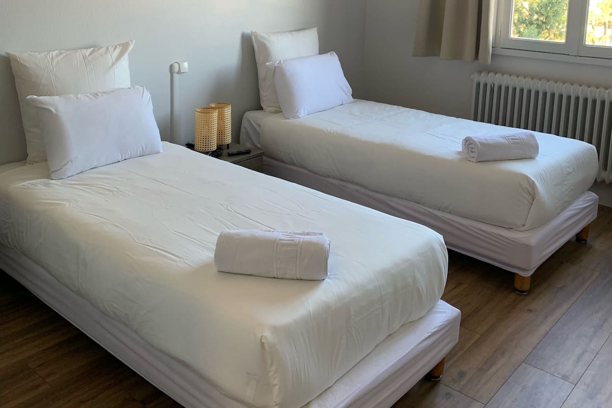 Quarto com duas camas de solteiro, cada uma com roupa de cama e almofadas brancas, mesa de cabeceira com luminária, e janela com cortinas.