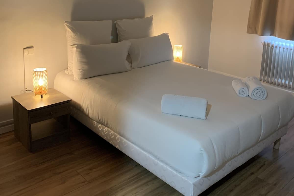 Uma cama bem arrumada com lençóis e almofadas brancos, uma luminária de cabeceira e uma toalha enrolada sobre a cama.
