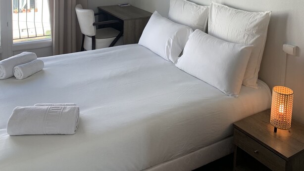 A hotel room with a large bed, a desk, and a chair. There is a window with curtains, a nightstand with a lamp, and a view of the outside. - Le Costellan (Dagneux)