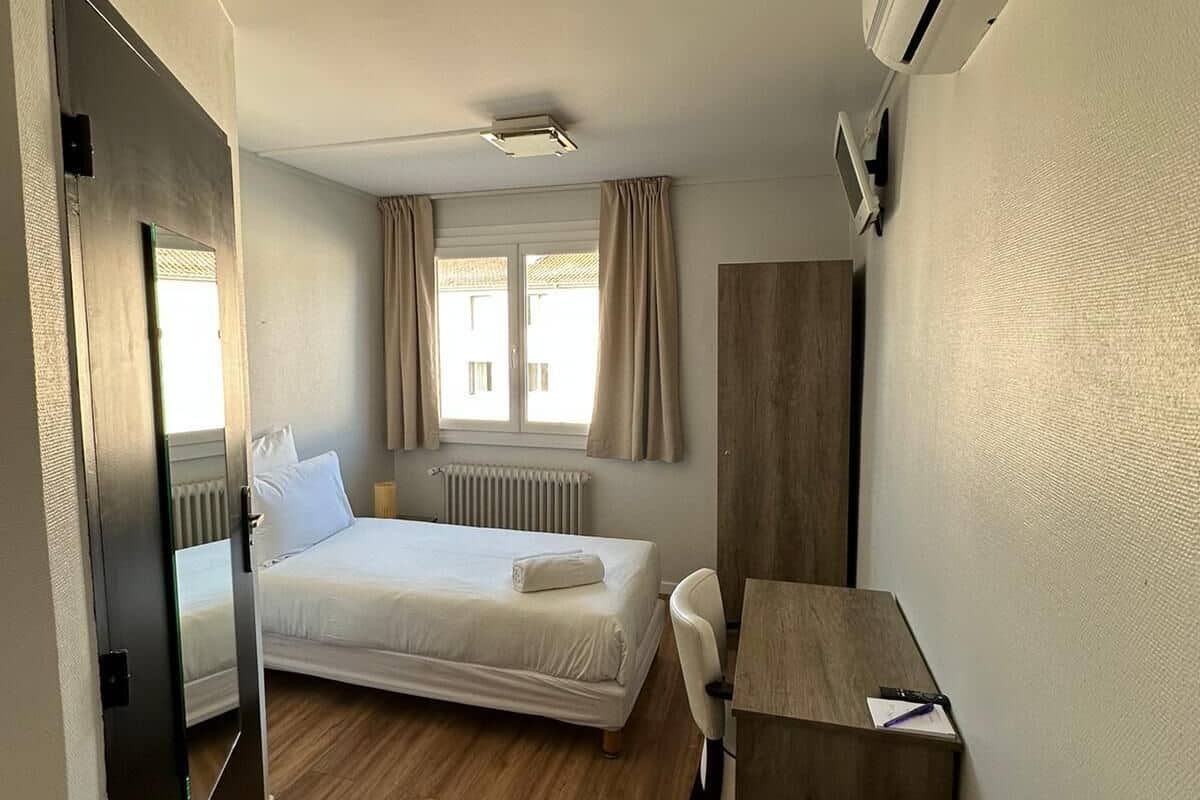 Quarto de hotel compacto com uma cama de solteiro, uma escrivaninha e um aparelho de ar condicionado.
