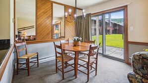 Un coin repas avec une table et des chaises en bois, un lustre et une vue sur un bâtiment et une pelouse, visible à travers une porte vitrée.