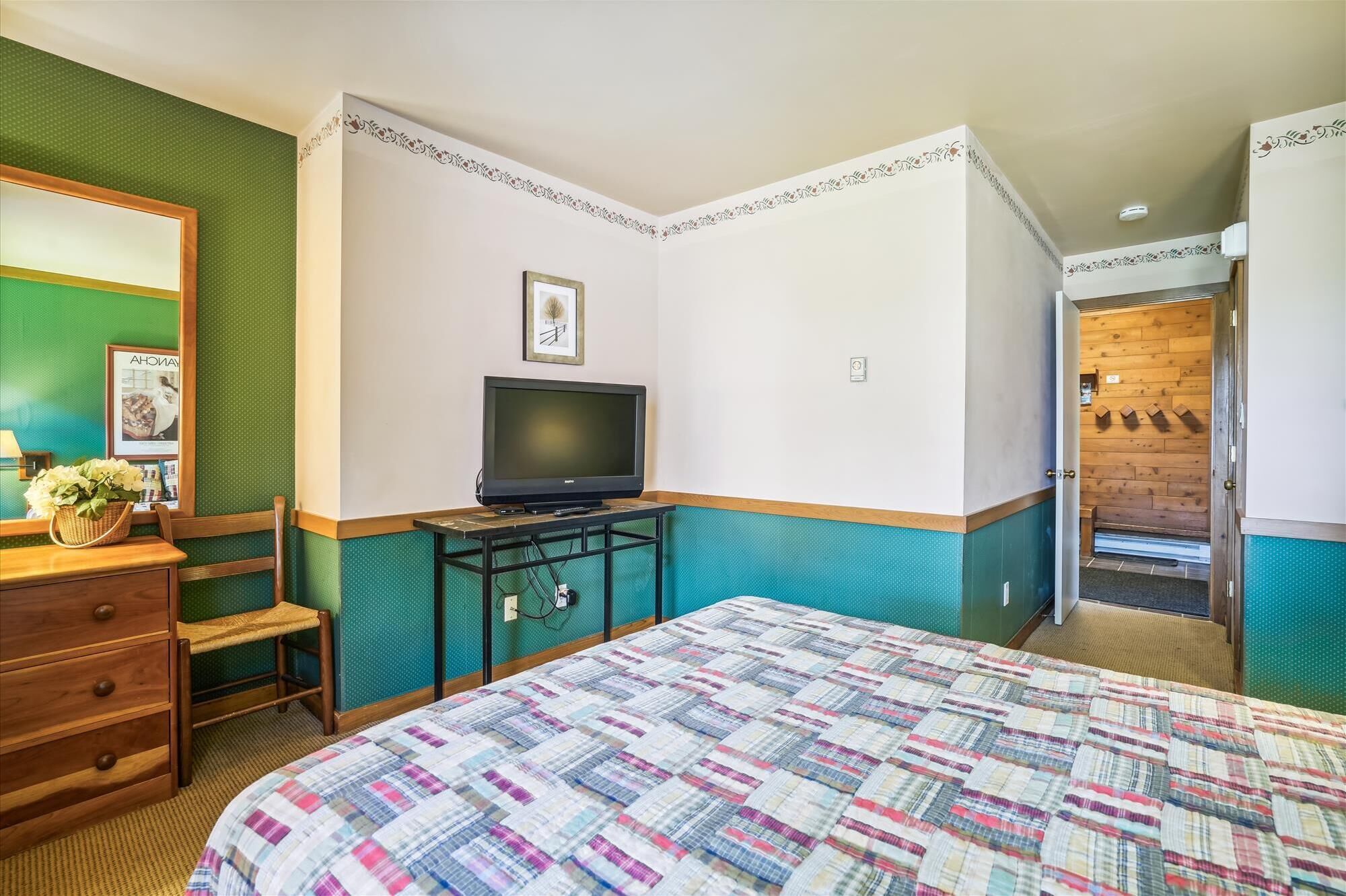A hotel room with a bed, a desk with a television, a dresser, and a wall with a decorative border.