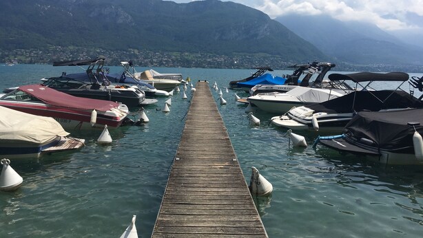 A wooden pier leading to a dock with various boats, a mountainous backdrop, and a clear sky. - Pause Café (Annecy)