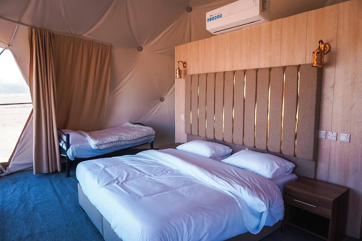 A tented room with two beds, a wooden headboard, an air conditioning unit, and a small bedside table.