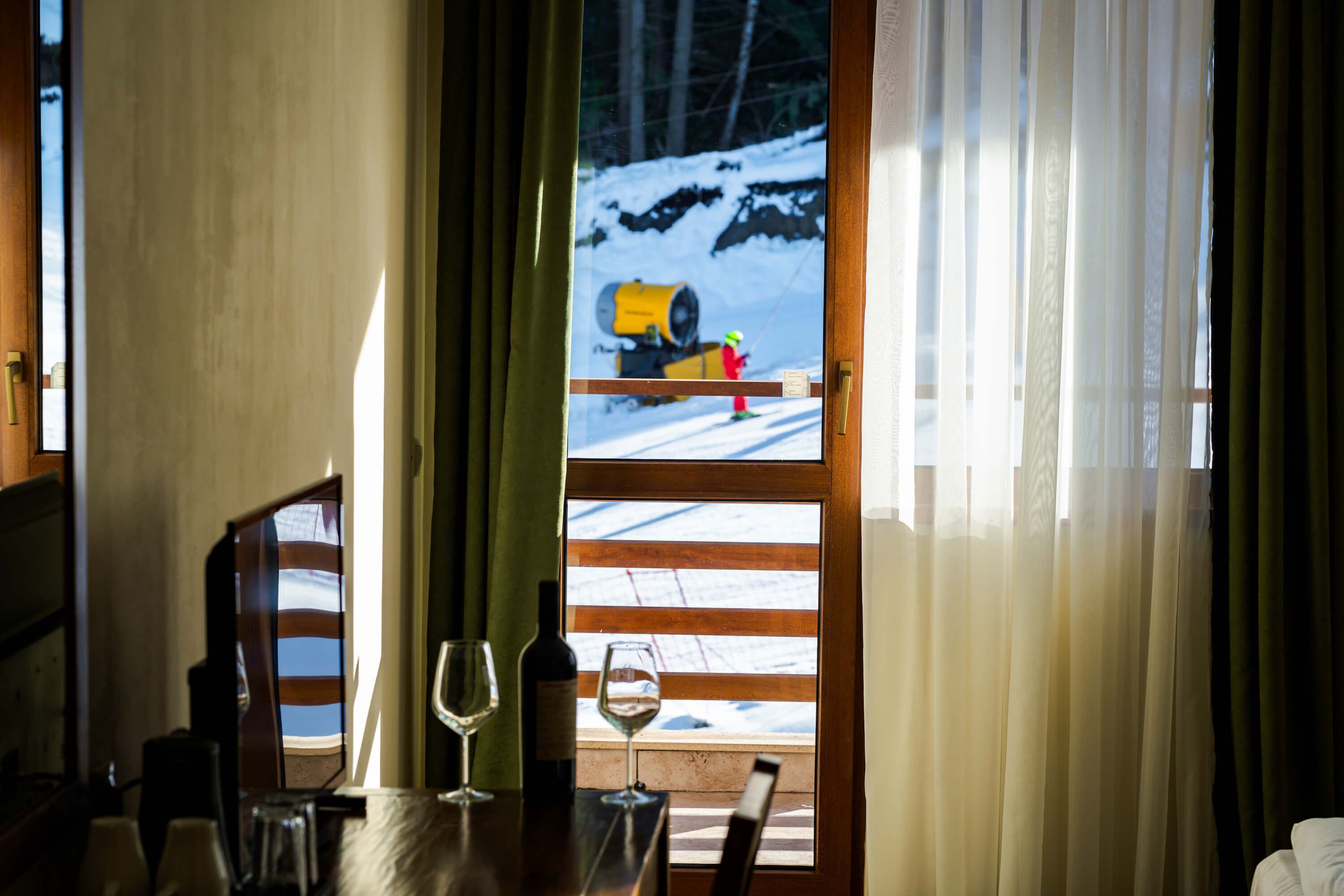 A room with a view of a snowy outdoor area, a wooden table with wine glasses, and a window with curtains.