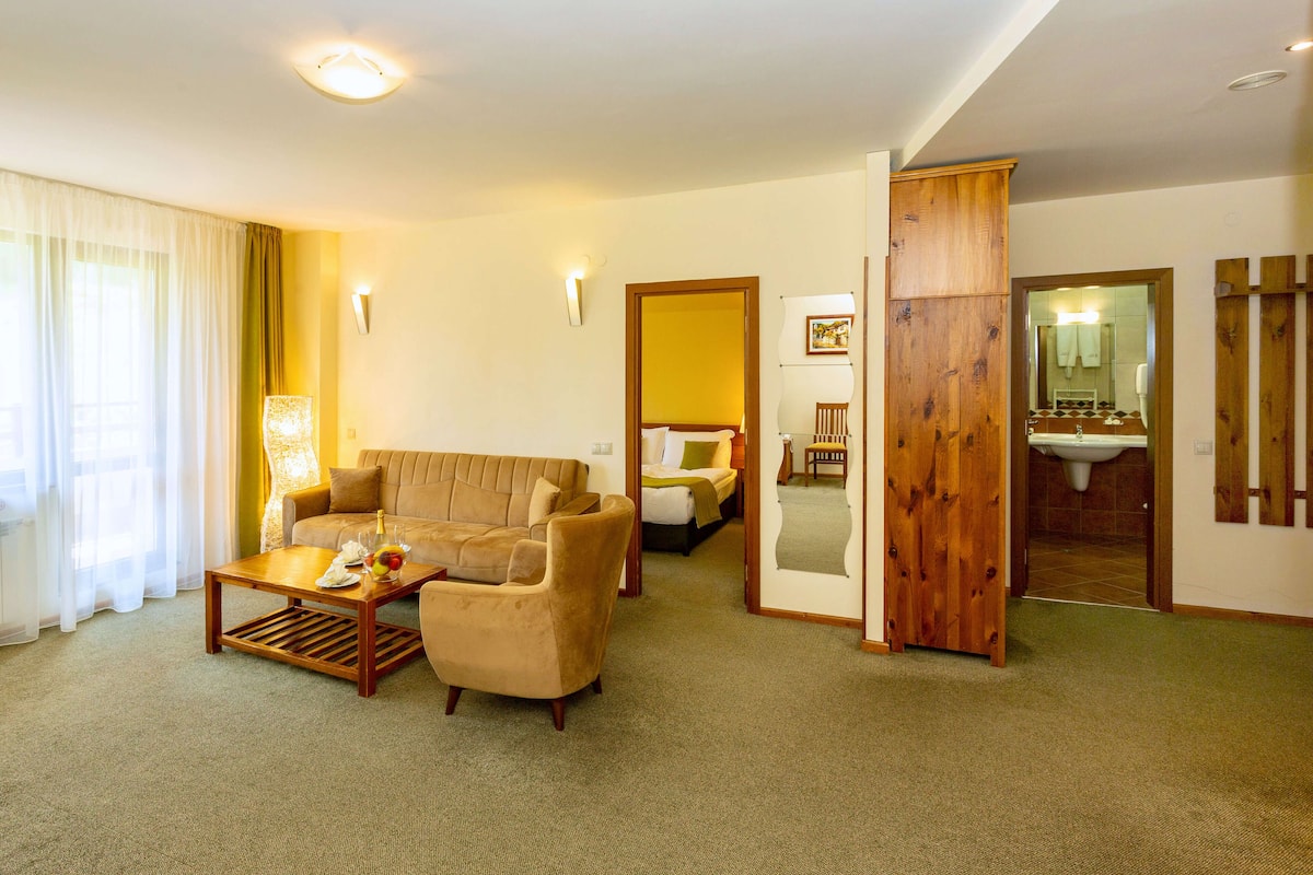Habitación de hotel con sala de estar que incluye un sofá, una mesa de centro y una lámpara. Hay una puerta de madera que da a un baño con lavamanos y espejo.