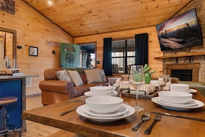 A wooden interior with a dining table set for four, a leather sofa, a fireplace, and a painting on the wall.