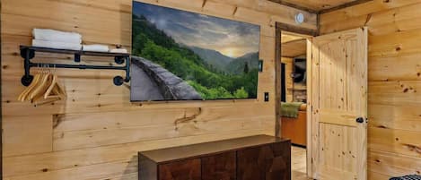 A wooden cabin interior with a large painting of a landscape, a wooden bench, and a door leading to another room.