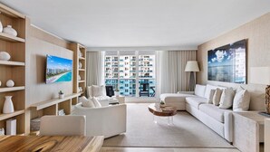Ein modernes Wohnzimmer mit Sofa, Couchtisch und Fernseher. Regale mit Dekorationselementen und ein großes Fenster mit Blick auf ein Gebäude.