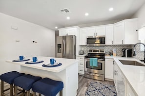 A modern kitchen with white cabinets, stainless steel appliances, a dining area with blue stools, and a patterned rug. - Tranquil Casa - Sparkling Pool & Outdoor Entertaining (Phoenix)