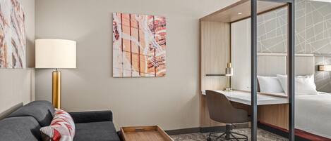 A hotel room with a grey sofa, a wooden side table, a desk, and a bed with a headboard.