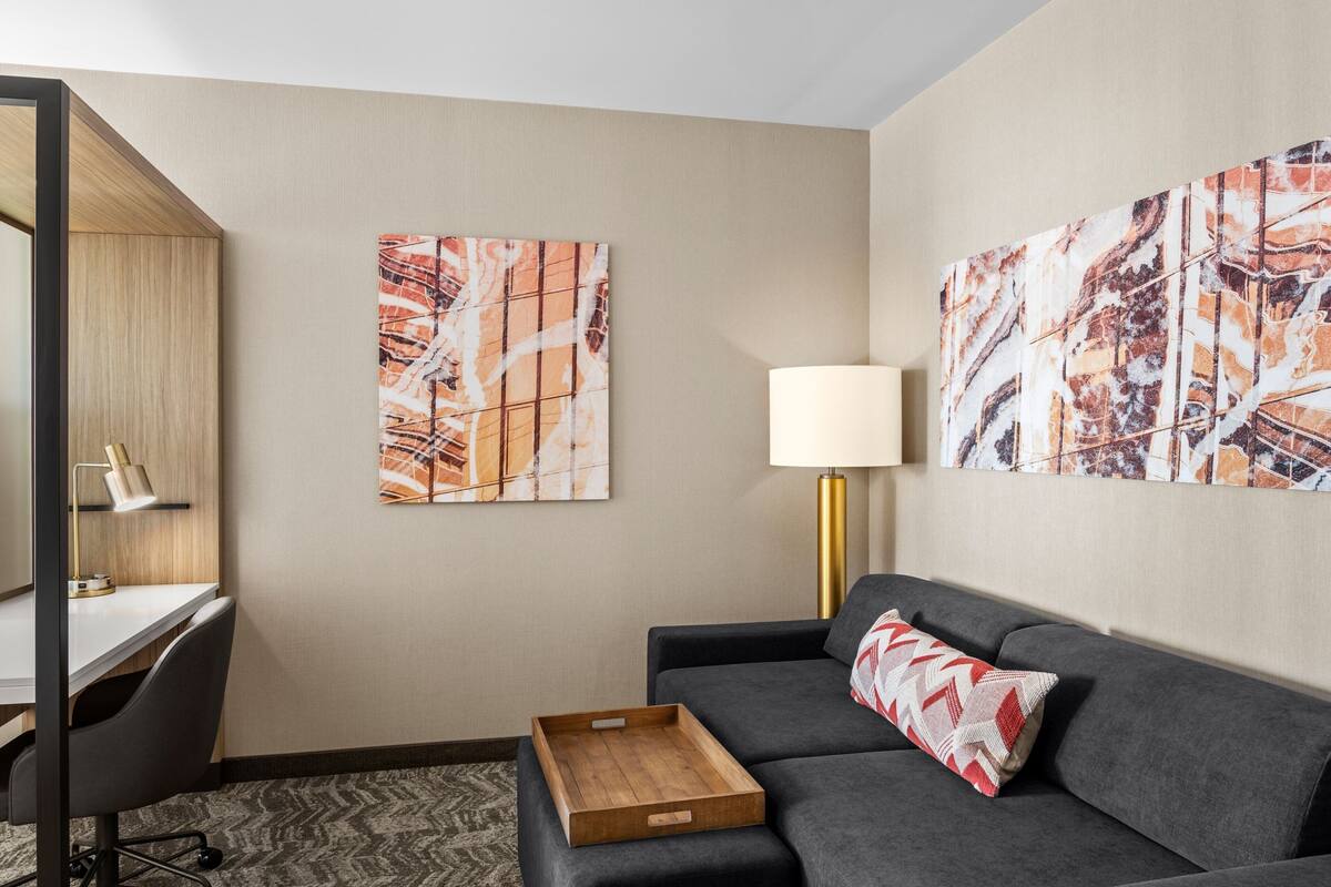 Une chambre d’hôtel moderne, dotée d’un bureau, d’un canapé gris foncé et de deux tableaux abstraits accrochés aux murs.