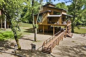 A two-story wooden cabin with a deck, surrounded by trees and a body of water.