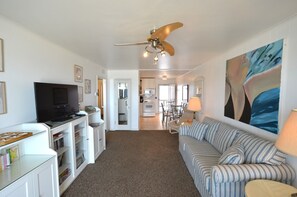 A living room with a television, bookshelves, a sofa, and a ceiling fan. - Lovely Ocean Front Condo In The Beachcomber! (Dewey Beach)
