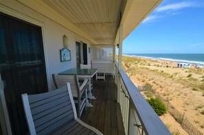 Une terrasse en bois avec une table et des chaises, donnant sur une plage bordée de dunes de sable et l’océan.