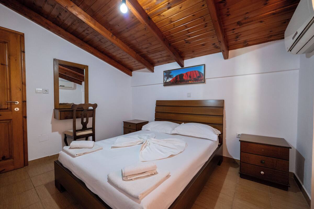 A bedroom with a wooden ceiling, a bed with white linens, a wooden nightstand, a wooden dresser, and a painting on the wall.