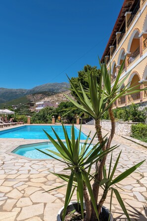 A pool area with a palm tree, stone tiles, and a building with balconies in the background. - Apart Hotel Alex (Himarë)