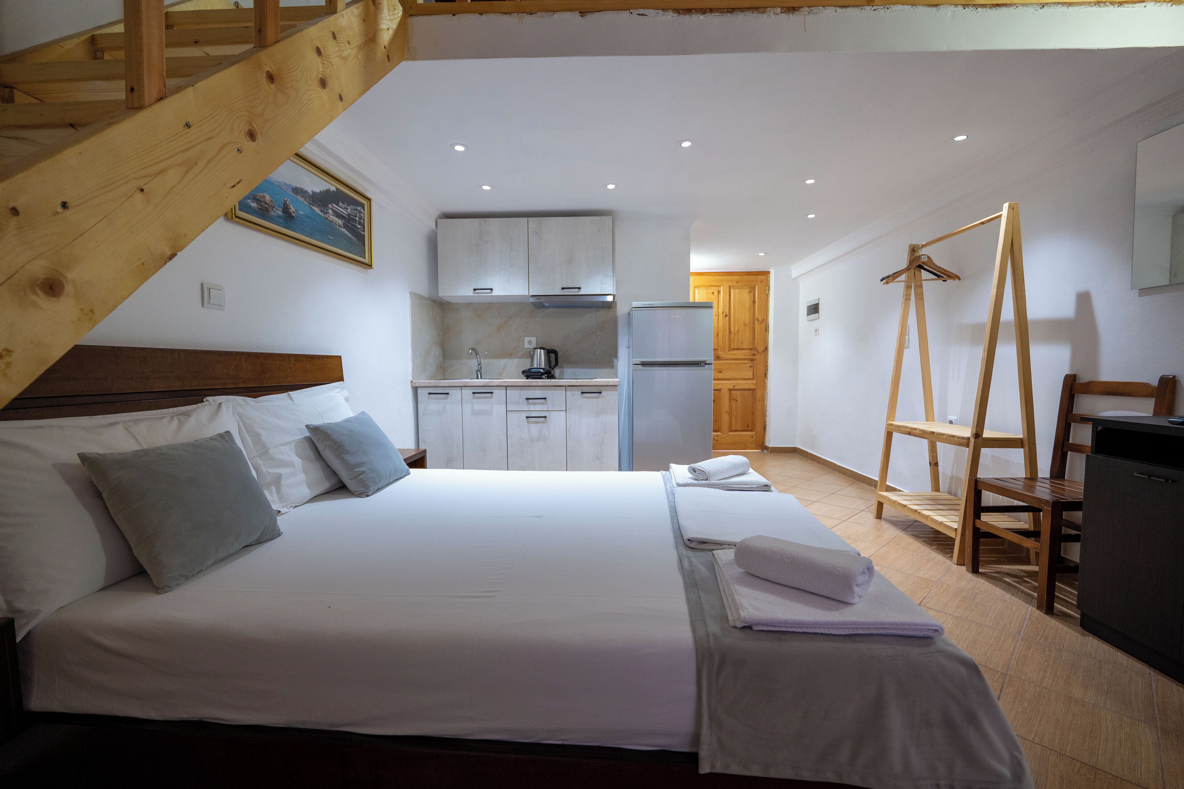 Een slaapkamer met een bed, kussens, een houten trap, een keukenhoek en een houten stoel.