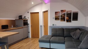 Una cucina moderna con mobili in legno, forno integrato e piano di lavoro in legno. Si nota un divano nero con un cuscino grigio e un piccolo tavolo con uno sgabello. La stanza presenta un pavimento in legno e un soffitto bianco con una striscia di luce viola.