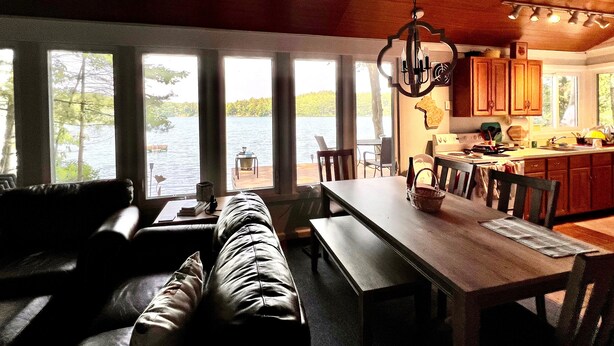 A cozy living room with a wooden ceiling, large windows overlooking a lake, a dining area with a wooden table and chairs, and a kitchen with wooden cabinets.