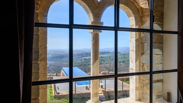 Vista a través de una ventana con arcos de piedra, una piscina y un paisaje lejano.