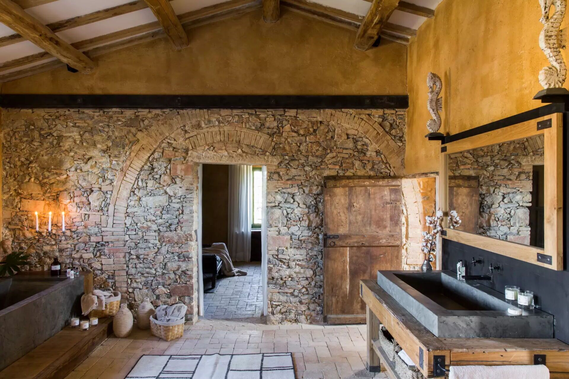 A rustic bathroom with a stone archway, wooden beams, a large mirror, and a stone sink.