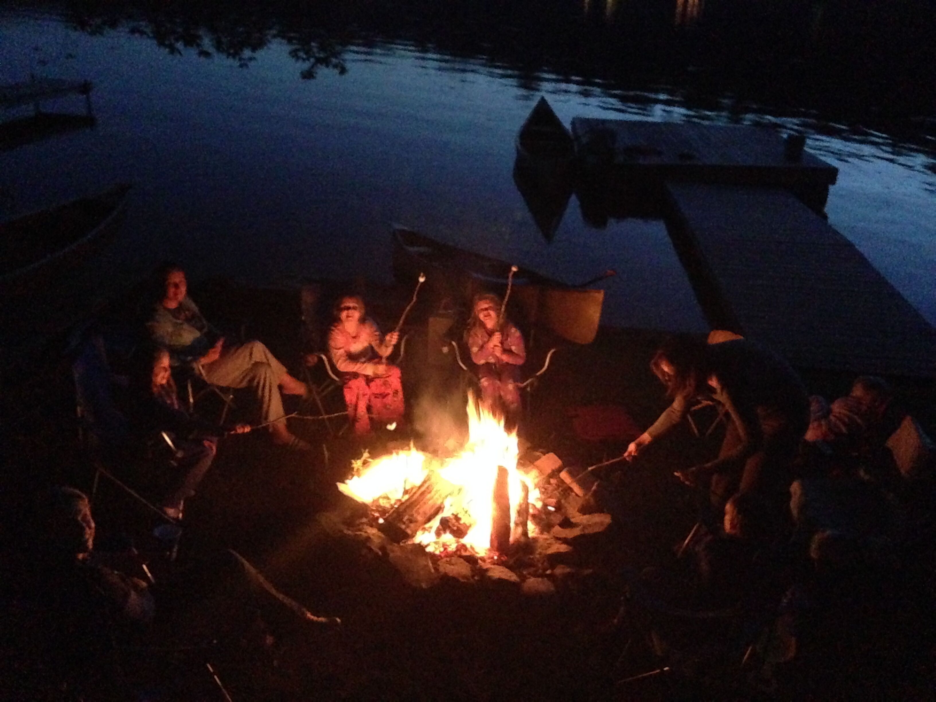 A group of people gathered around a campfire on a dock by a body of water at night.