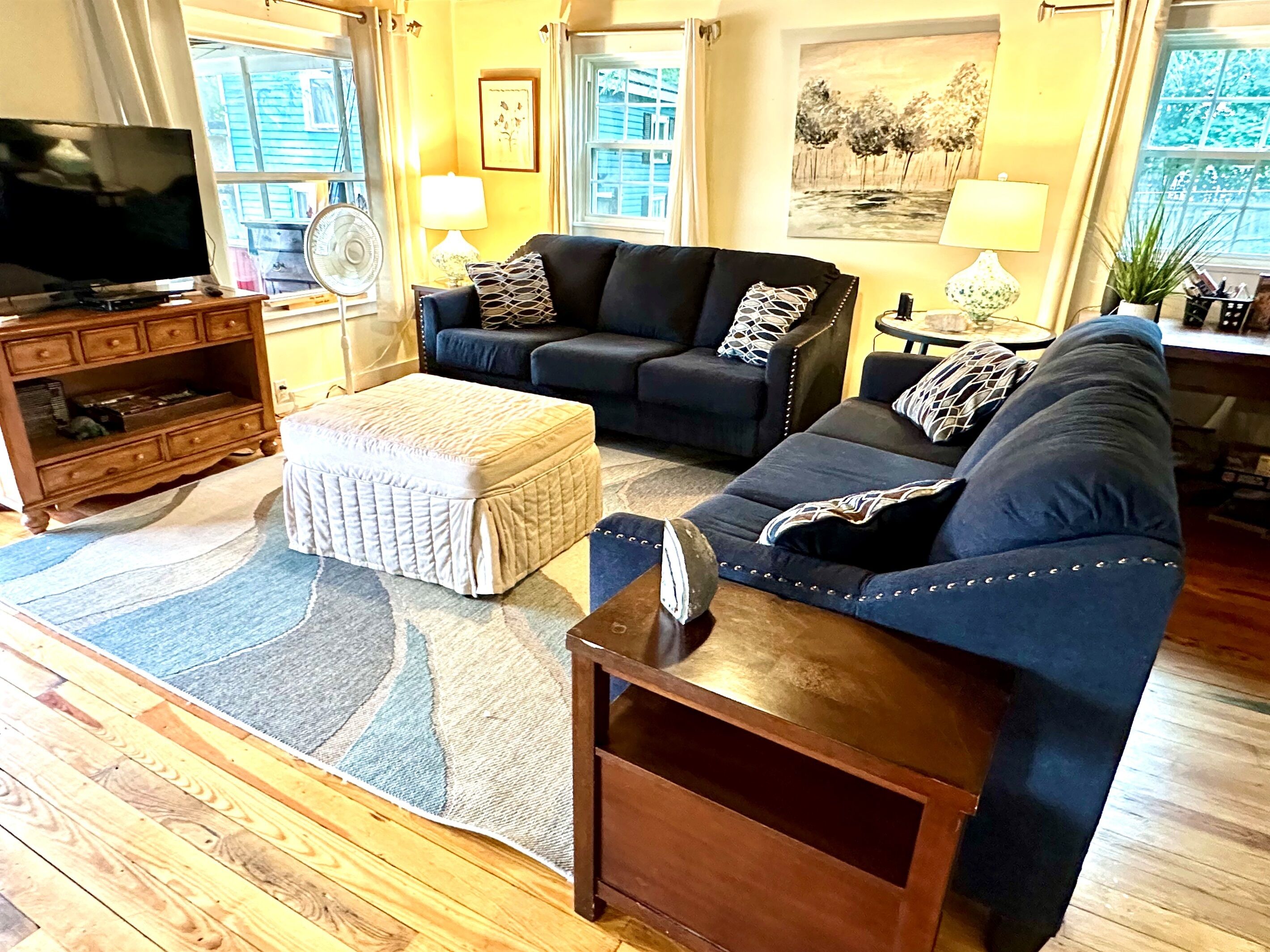 A living room with a sofa set, ottoman, wooden floor, and a television on a wooden cabinet.