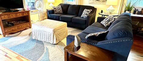 A living room with a sofa set, ottoman, wooden floor, and a television on a wooden cabinet.