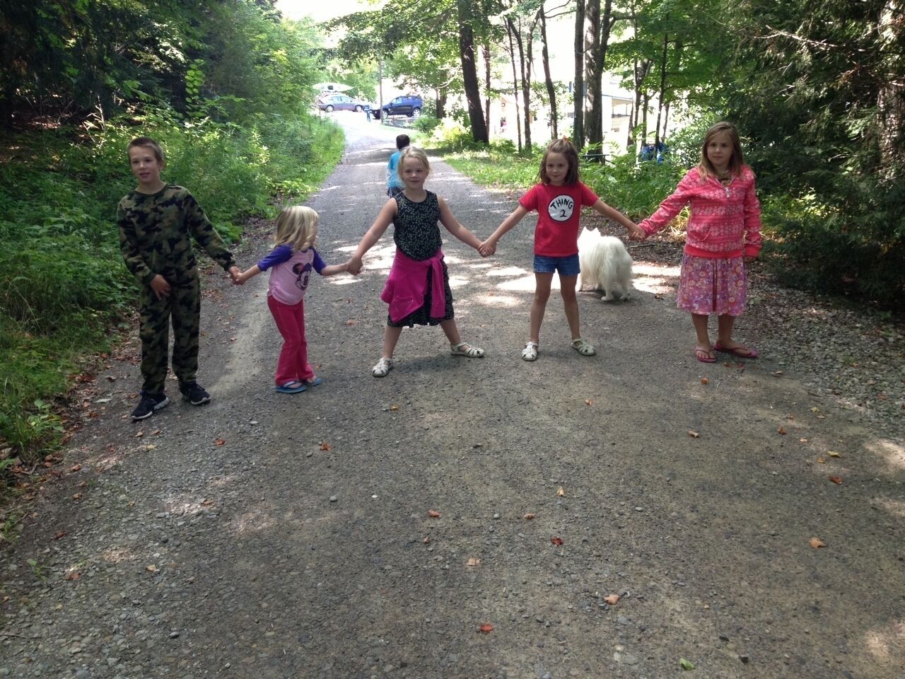 A group of children holding hands on a dirt path surrounded by trees.