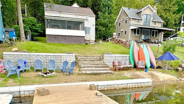 A wooden dock leading to a calm water body, surrounded by a stone wall and a grassy area with blue Adirondack chairs. - Lake Demmon Lodge-Beach,Boats,Fishing,FUN!! (Bath)