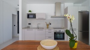 Una cocina moderna con armarios blancos, un refrigerador de acero inoxidable, un microondas y un horno. Hay una mesa de madera con un mantel tejido y un tazón.