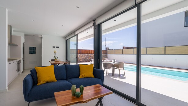 Un moderno salón con un sofá azul, una mesa de centro de madera con objetos decorativos y vistas a una piscina a través de puertas de vidrio grandes.