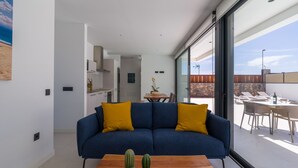 Una sala de estar moderna con un sofá azul, una mesa de centro de madera con cactus y un gran ventanal corredero que da a un patio exterior con mesa y sillas.