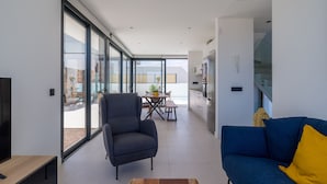 En moderne stue med sofa, lænestol og sofabord, et køkkenområde med køleskab og skabe, samt store skydedøre til en udendørs terrasse.